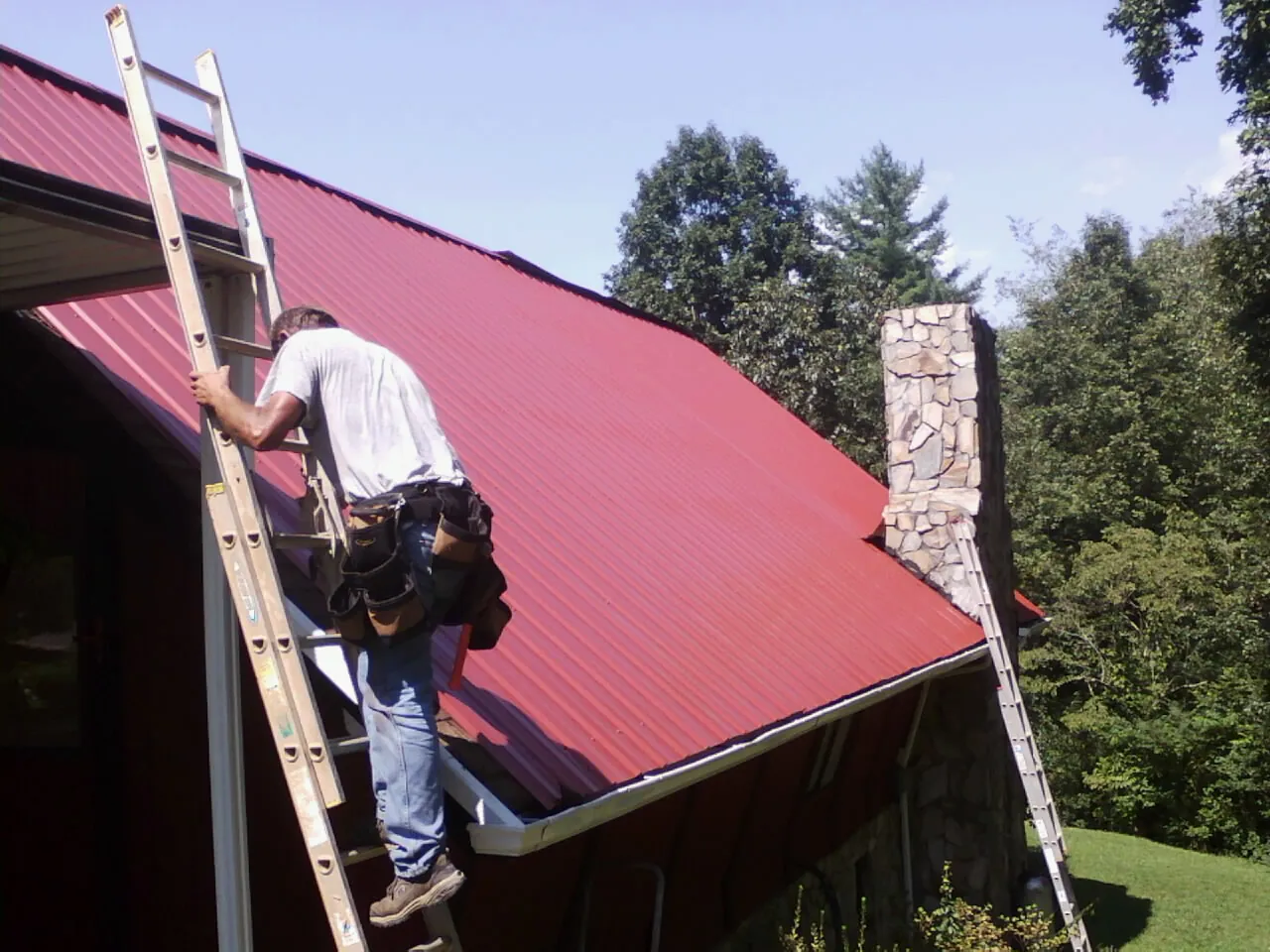 Roofer on a ladder beside a red metal roof and stone chimney — Soffit & Fascia Repair by Roofing Brothers in Achille